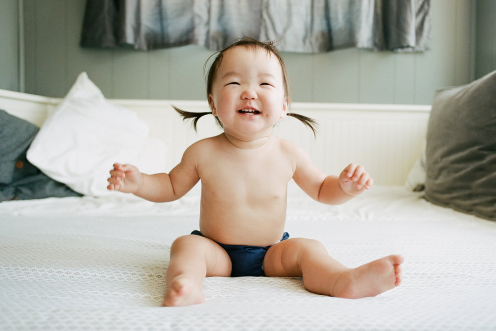 Family photography: A laughing baby sitting on a bed.