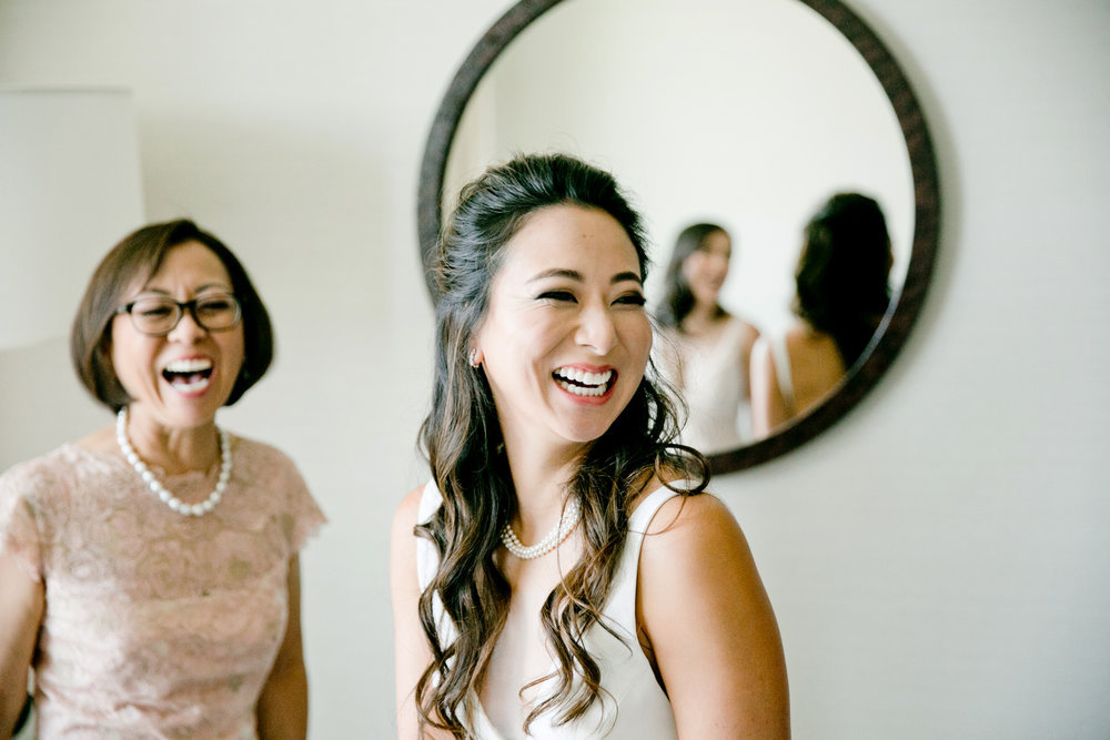 A bride and her mother laughing in front of a mirror.