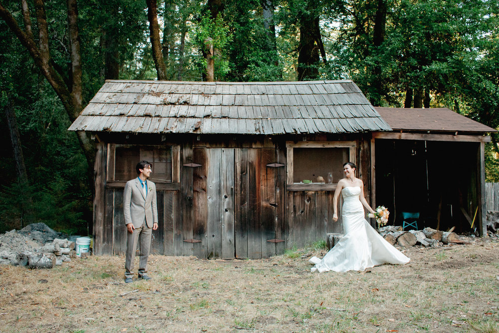 A bride and groom standing in front of a wooden shack.