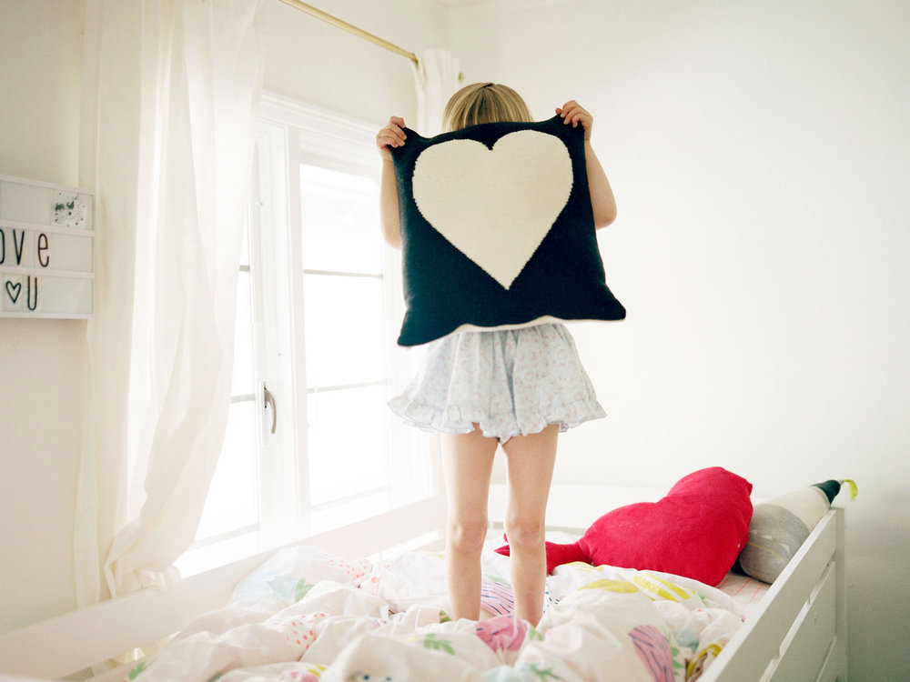 young girl standing on her bed holding heart pillow over her face playfully