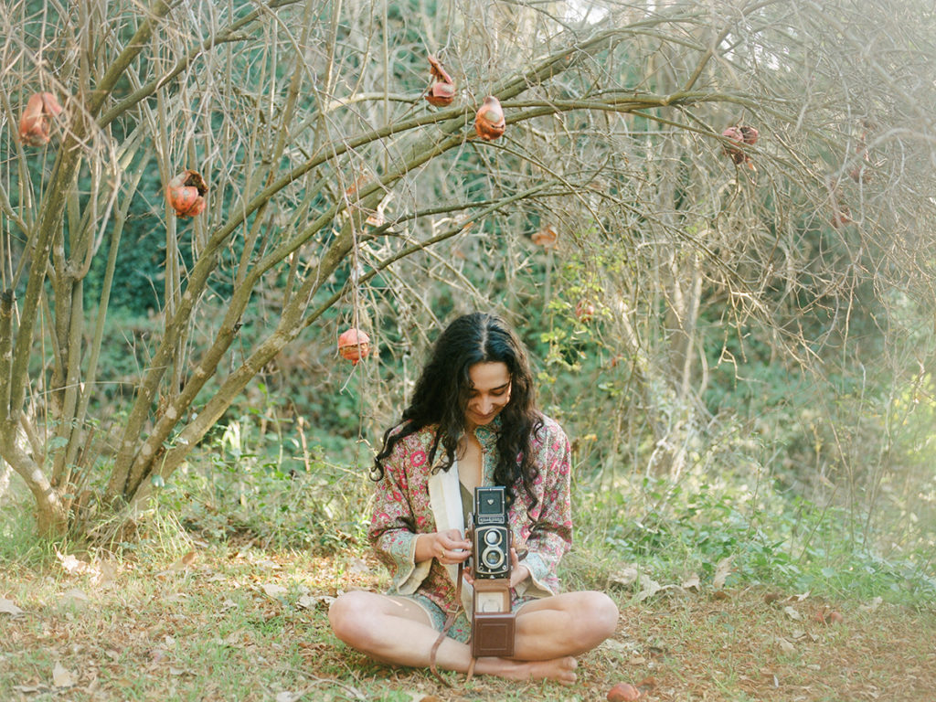woman sitting cross legged under a tree and looking into a vintage camera