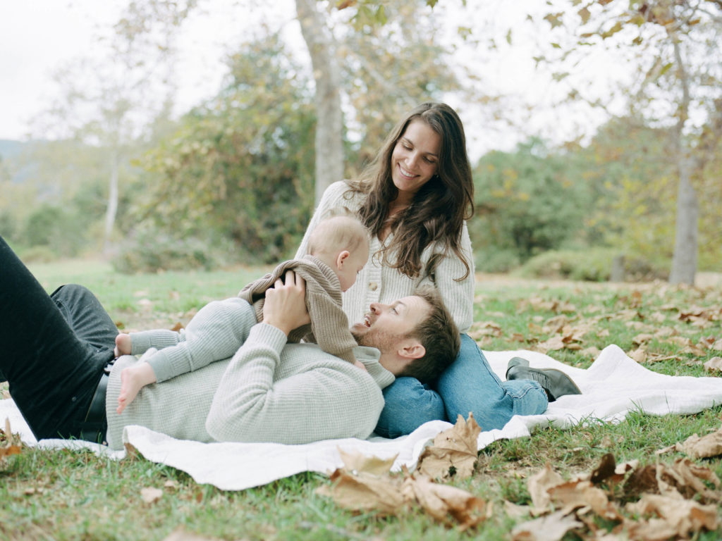 A family poses for a family photography session on a blanket in a park with their baby.