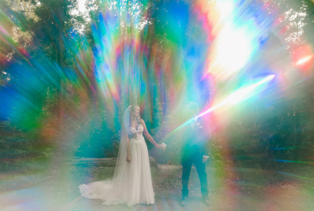 A bride and groom standing in front of a rainbow light beam.