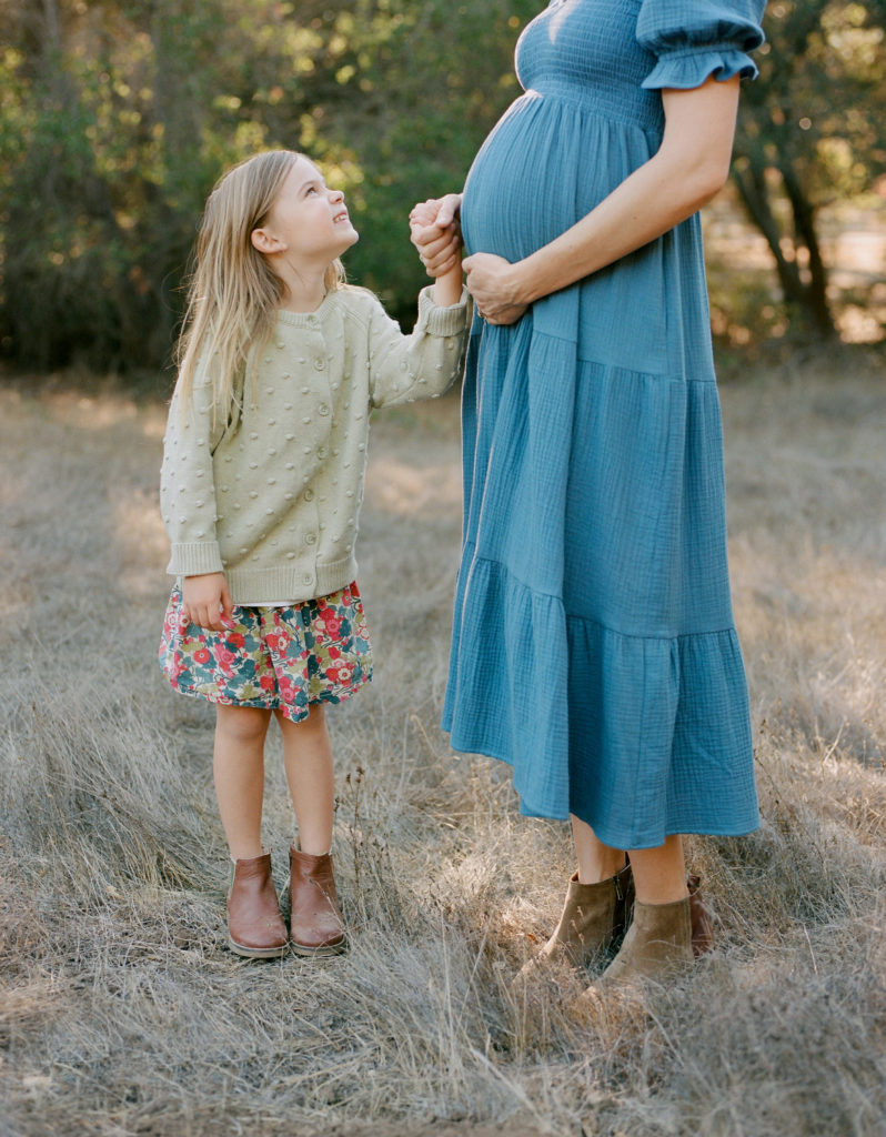 Family photography of a pregnant woman in a blue dress alongside a little girl in a field.