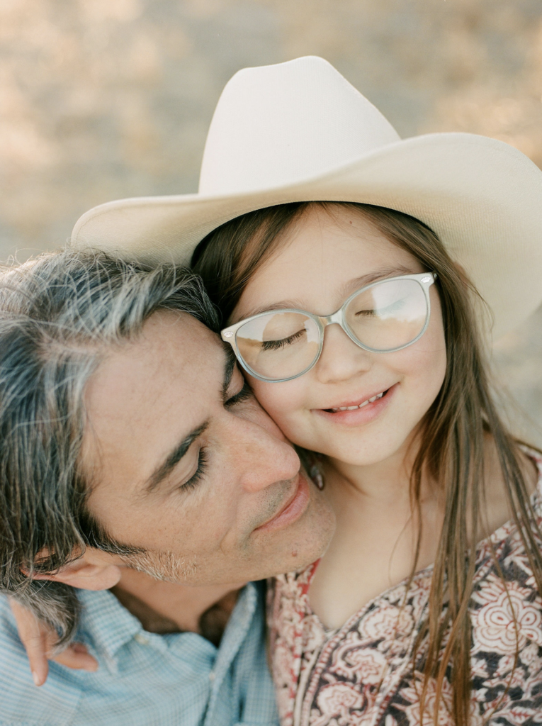 Family photography: A man affectionately embraces a young girl, adorned with glasses and a cowboy hat.