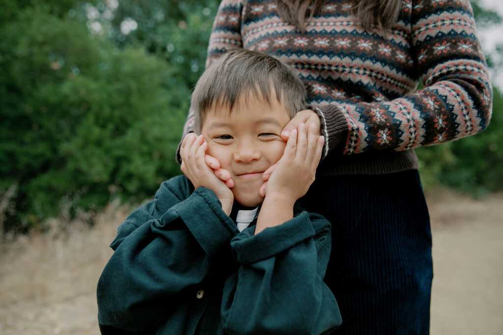 young boy smiling at camera with moms hands cupping cheeks from behind lovingly