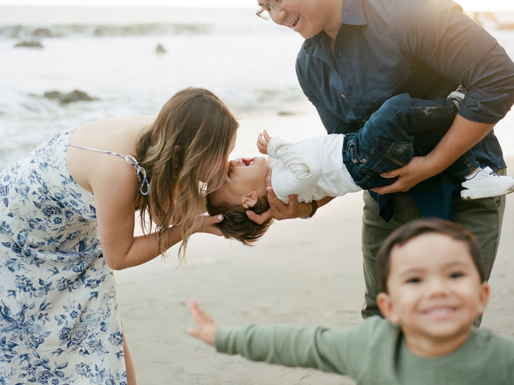 family playing at beach, mom kissing toddler who is upside down