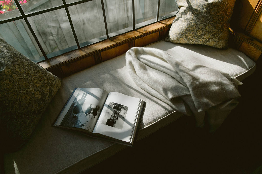 A cozy reading nook with sunlight filtering through the window, featuring a throw blanket and an open photo book on a bench.
