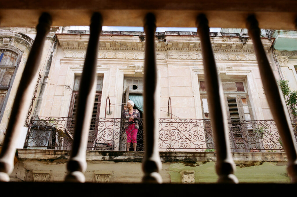 View from a balcony through iron bars showing another balcony with a person standing, old buildings in the background.