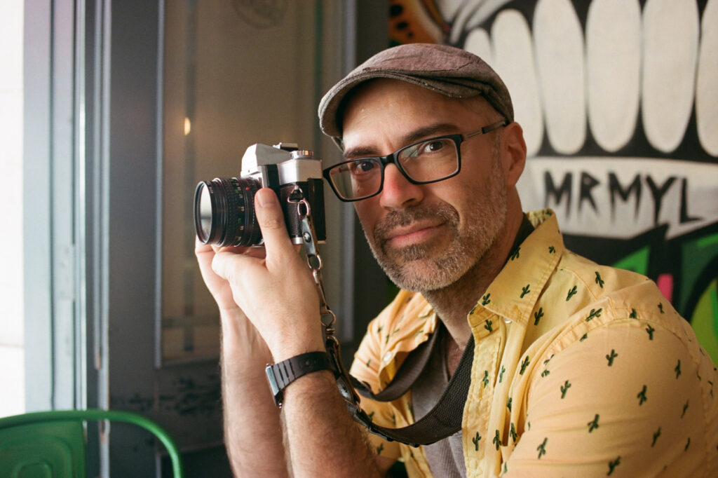 A man in a yellow shirt and cap holding a vintage camera.
