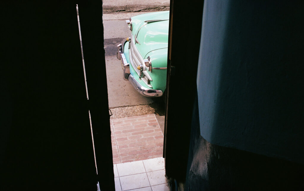 A vintage green car parked outside as viewed from a partially open doorway.
