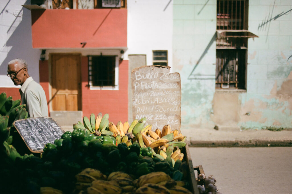 An elderly person walks past a street fruit stand with prices listed on a chalkboard in a sunny urban setting.