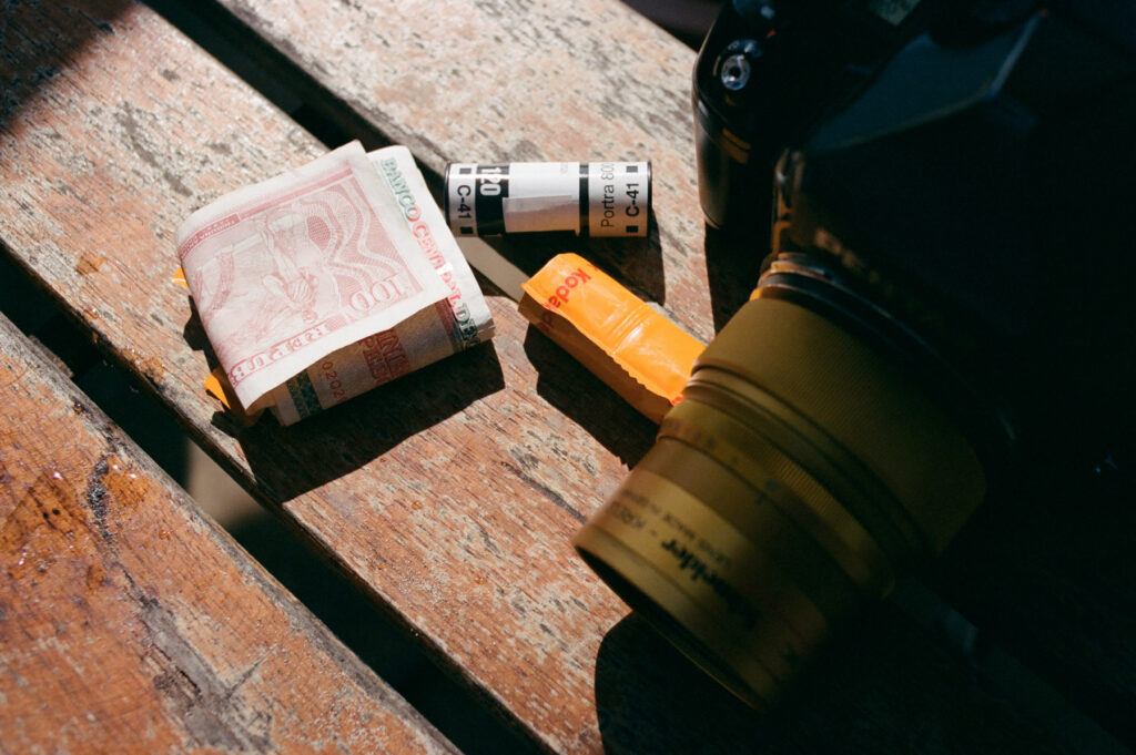 Photography essentials with a roll of film, currency, and a camera on a wooden surface bathed in sunlight.