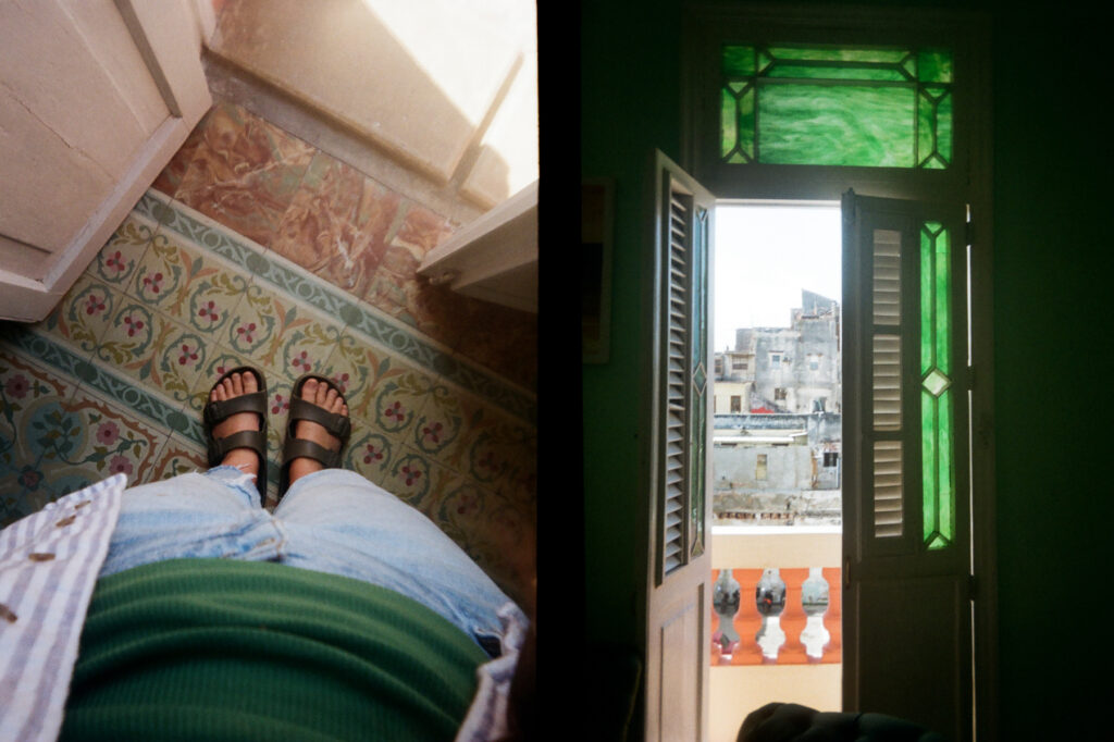 A split image showing a person's feet on a tiled floor in one half, and a view from a green shuttered window onto a cityscape in the other half.