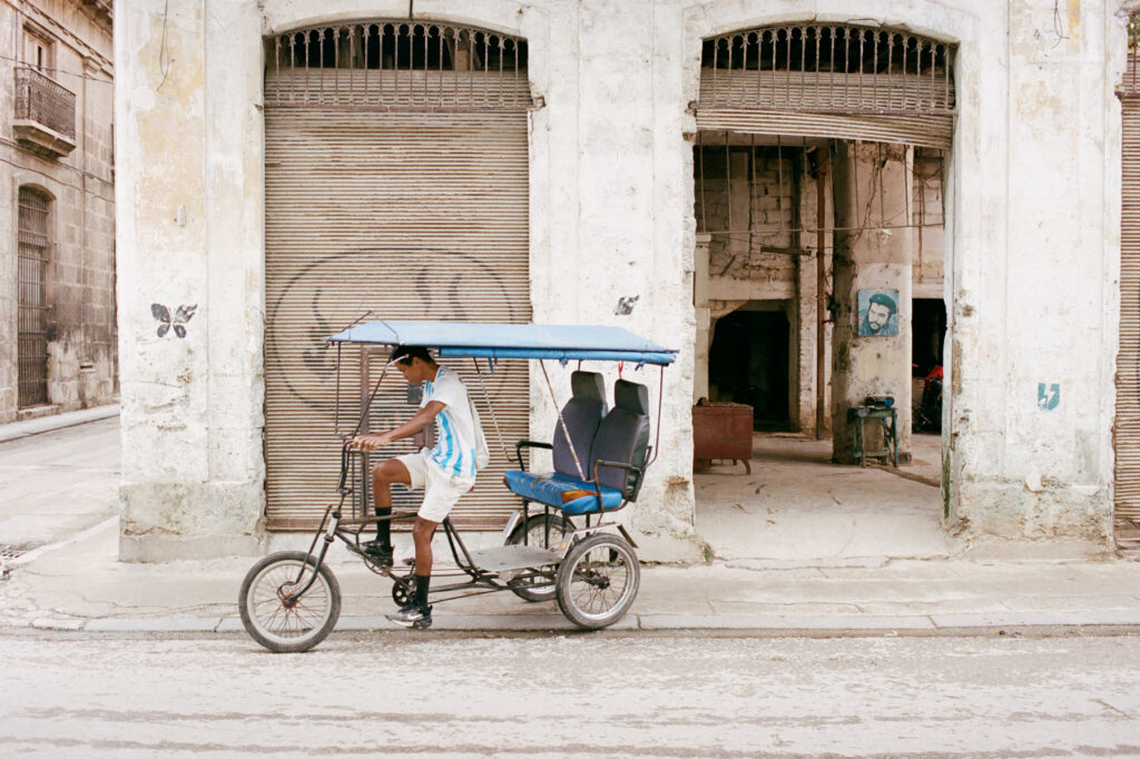 A person riding a bicycle taxi past weathered buildings with graffiti.
