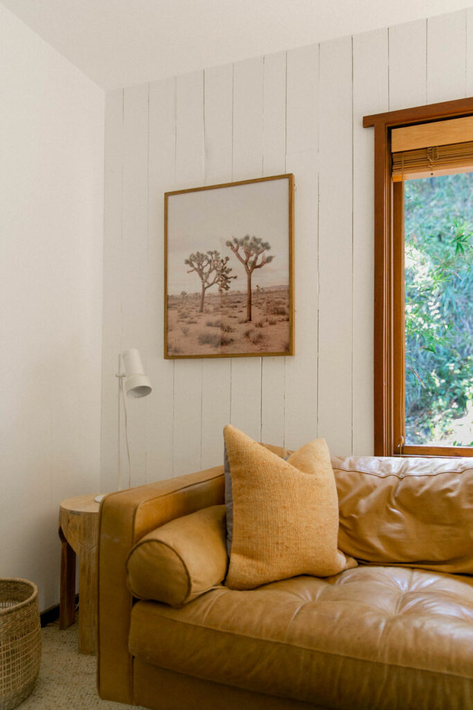 Cozy corner with a leather sofa, decorative pillows, and a framed desert landscape painting on a white paneled wall.