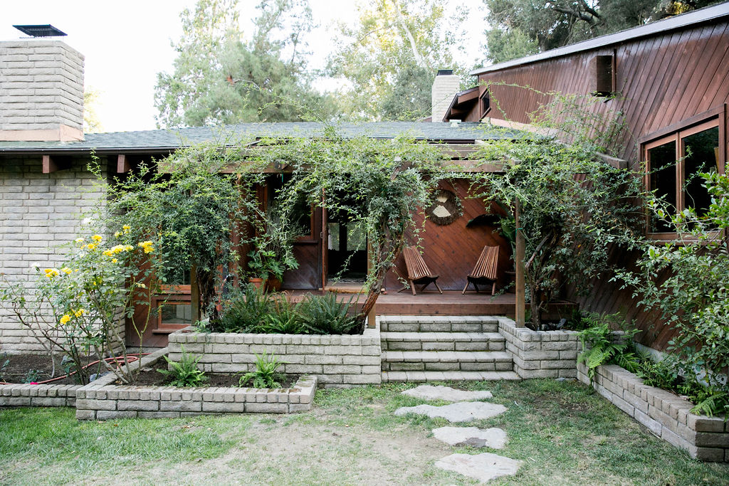 Plants overgrowing the entrance of a rustic house with a wooden facade and stepping stones leading to the porch.