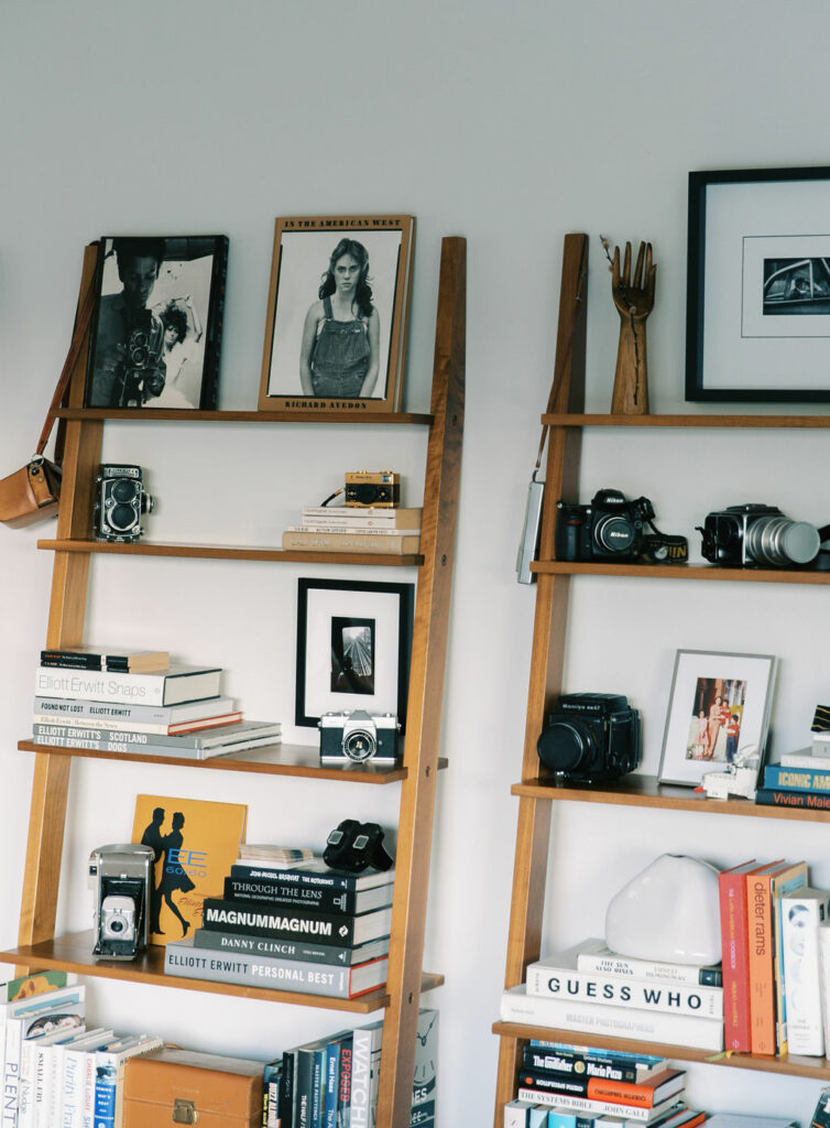 A wooden shelf displaying a collection of framed photographs, cameras, books, and decorative items against a light gray wall.