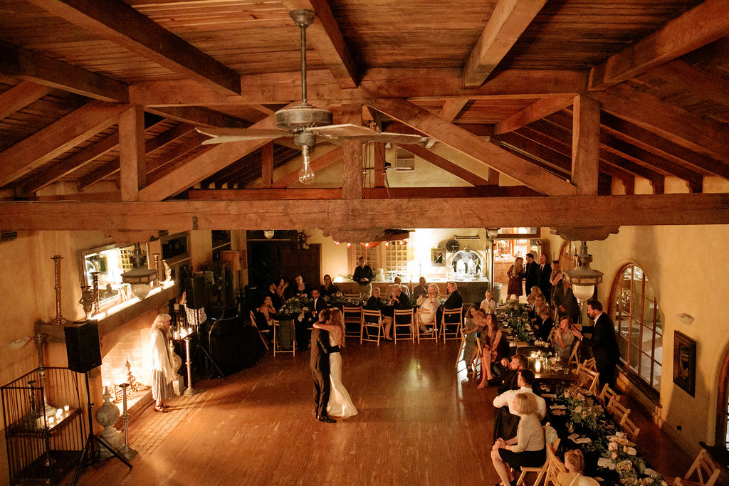 Warmly-lit rustic wedding reception with guests seated at tables and a couple in the center of the room.