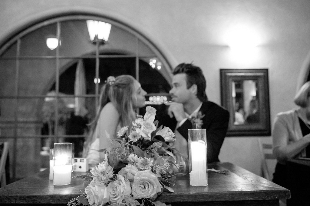A couple exchanges a tender gaze across a table adorned with flowers and candles at an indoor event.
