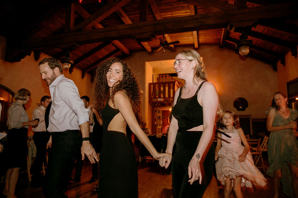 Two women holding hands and dancing with a smiling man at an indoor gathering.