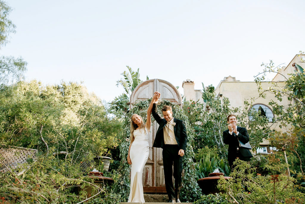 A newlywed couple holding hands and triumphantly raising their arms as they walk, with an applauding individual in the background, set in a garden with lush greenery.