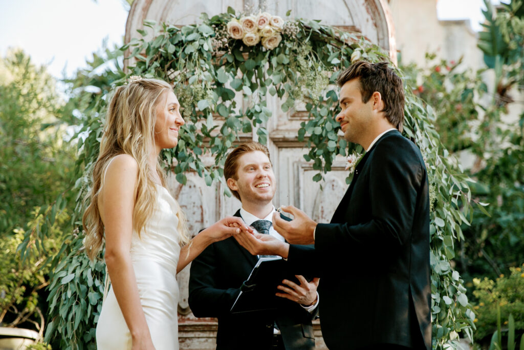 A bride and groom exchange vows in an outdoor ceremony officiated by a man with a book.