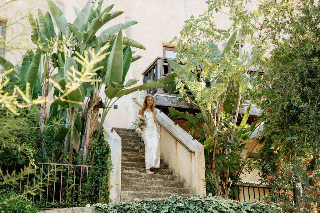 A bride descends an old stone staircase surrounded by lush greenery.