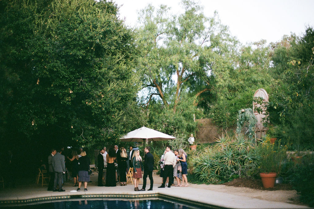 Group of people gathered by a pool in a garden setting for a social event.