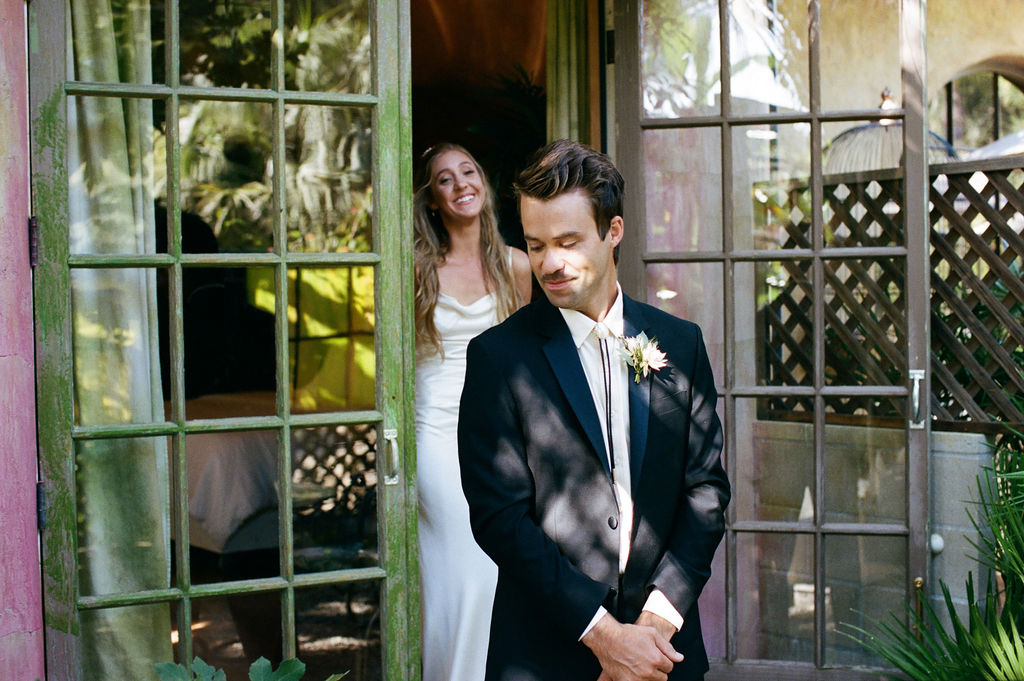 A bride happily looks on from a doorway as the groom smiles gently in the foreground.