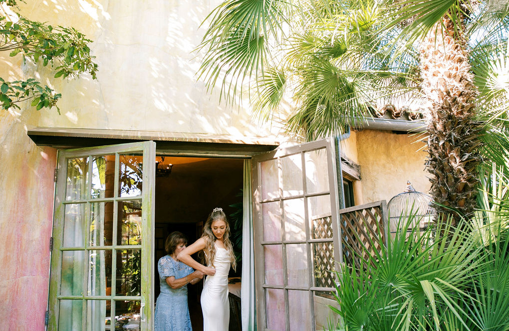 Two individuals embracing near an open door of an old house surrounded by lush greenery.