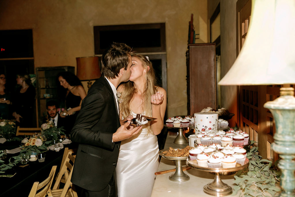 A couple shares a kiss while standing at a dessert table during a wedding reception.