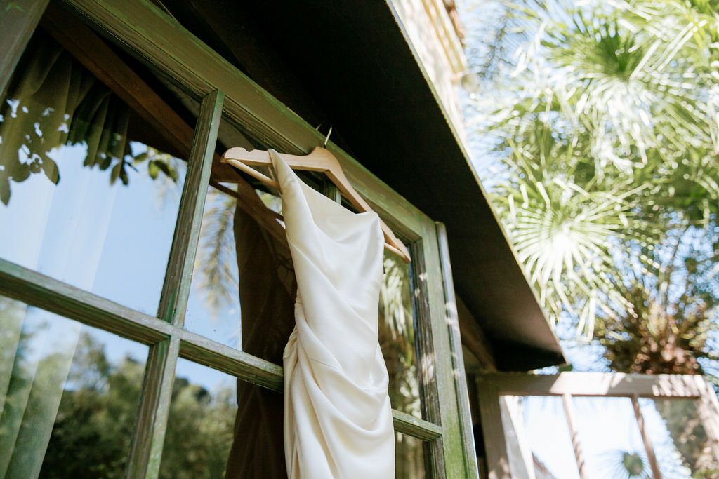 A wedding dress hanging on a window frame with greenery in the background.