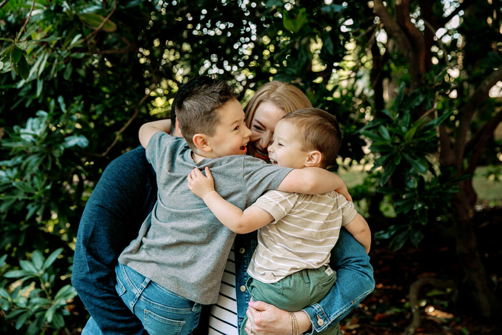 A woman embraces two young boys in a loving hug outdoors among greenery.
