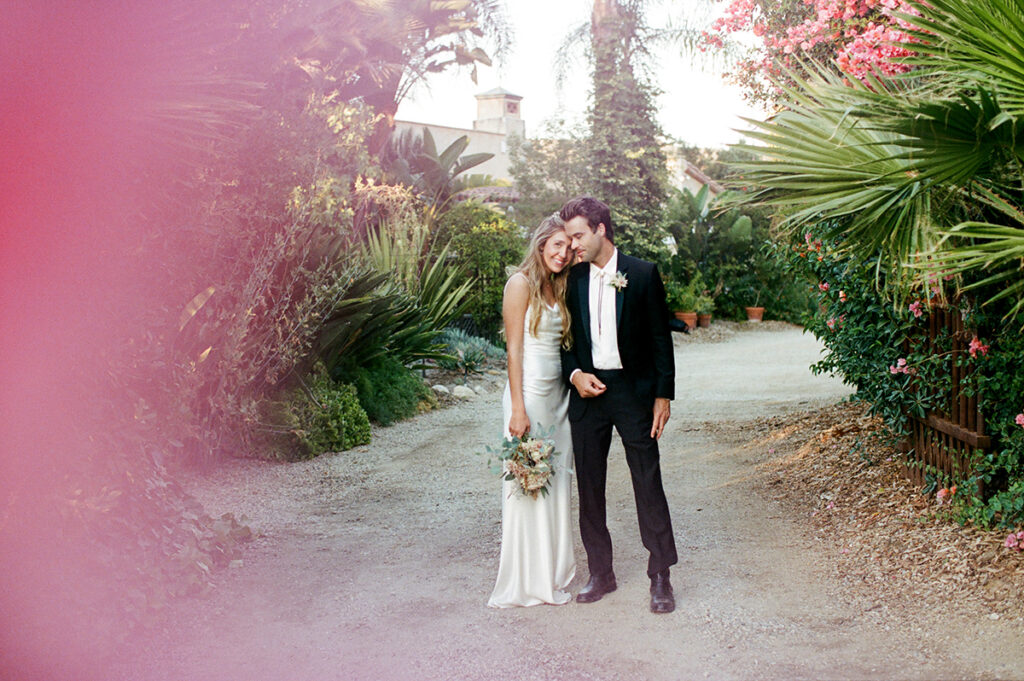 A couple in wedding attire lovingly embracing outdoors, with a warm, sunlit garden in the background.
