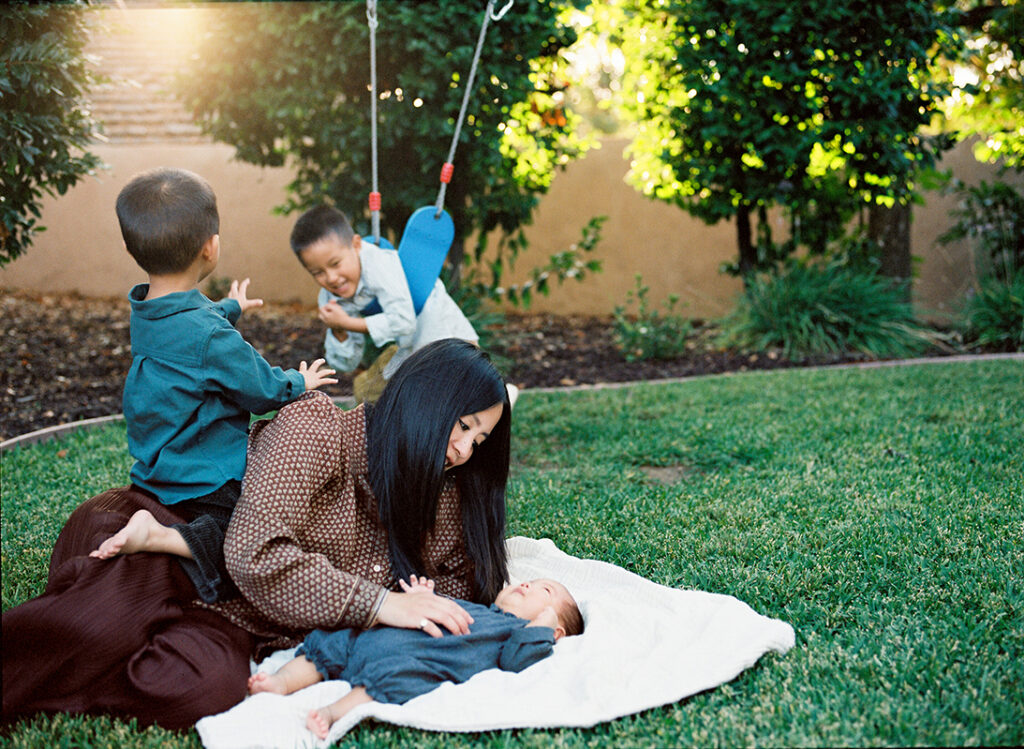 A family moment outdoors with a mother lying on the grass with a baby, as two other children play on a swing in the background.