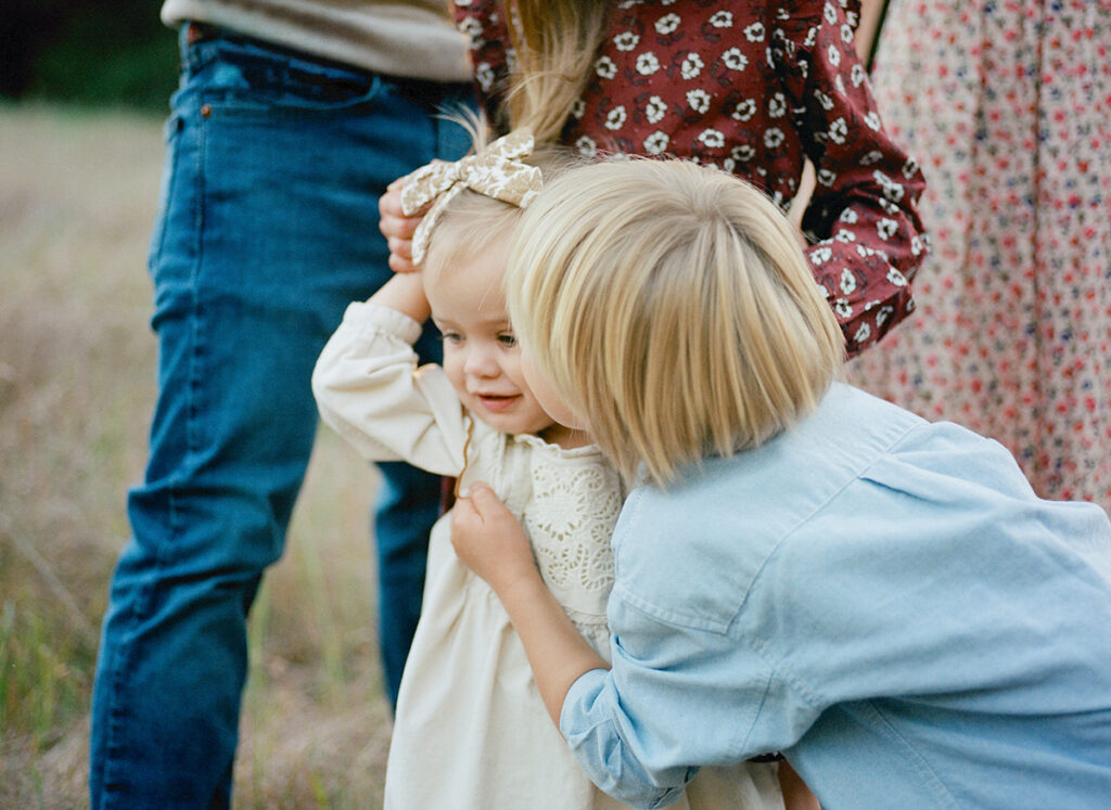 A child in a blue shirt gently embraces a younger child in a white dress outdoors, while two adults partially appear in the background.