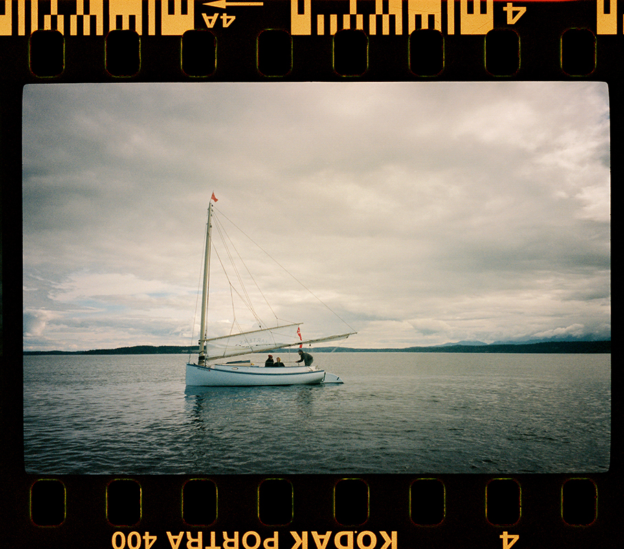 A sailboat with its sails down floating on calm waters under a cloudy sky, framed within the sprocket holes of a filmstrip.