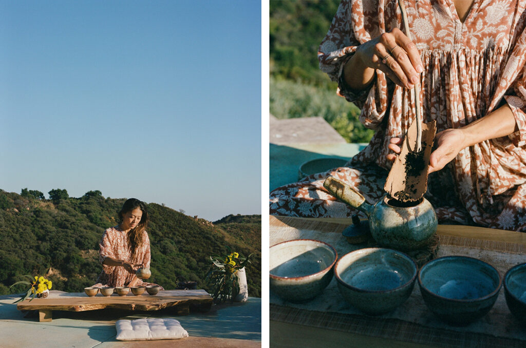 A woman prepares tea outdoors on a wooden table with ceramic bowls, surrounded by nature.