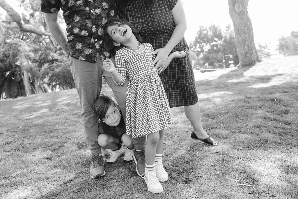A playful family moment outdoors with a young girl smiling and pointing upwards, another child crouching below, and the partial view of two adults.