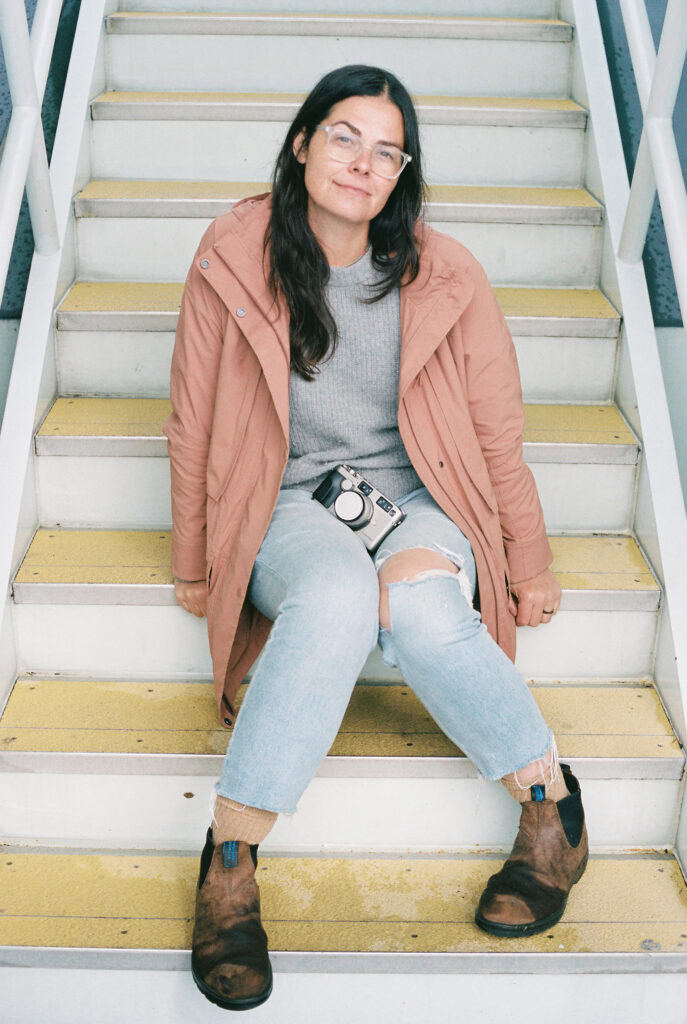 A person with long dark hair sits on stairs wearing glasses, a pink jacket, and jeans, with a camera resting on their lap.